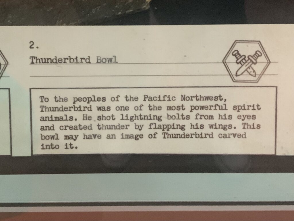 Museum label which reads '2. Thunderbird Bowl. To the peoples of the Pacific Northwest, Thunderbird was one of the most powerful spirit animals. He shot lightning bolts from his eyes and created thunder by flapping his wings. This bowl may have an image of Thunderbird carved into it.'